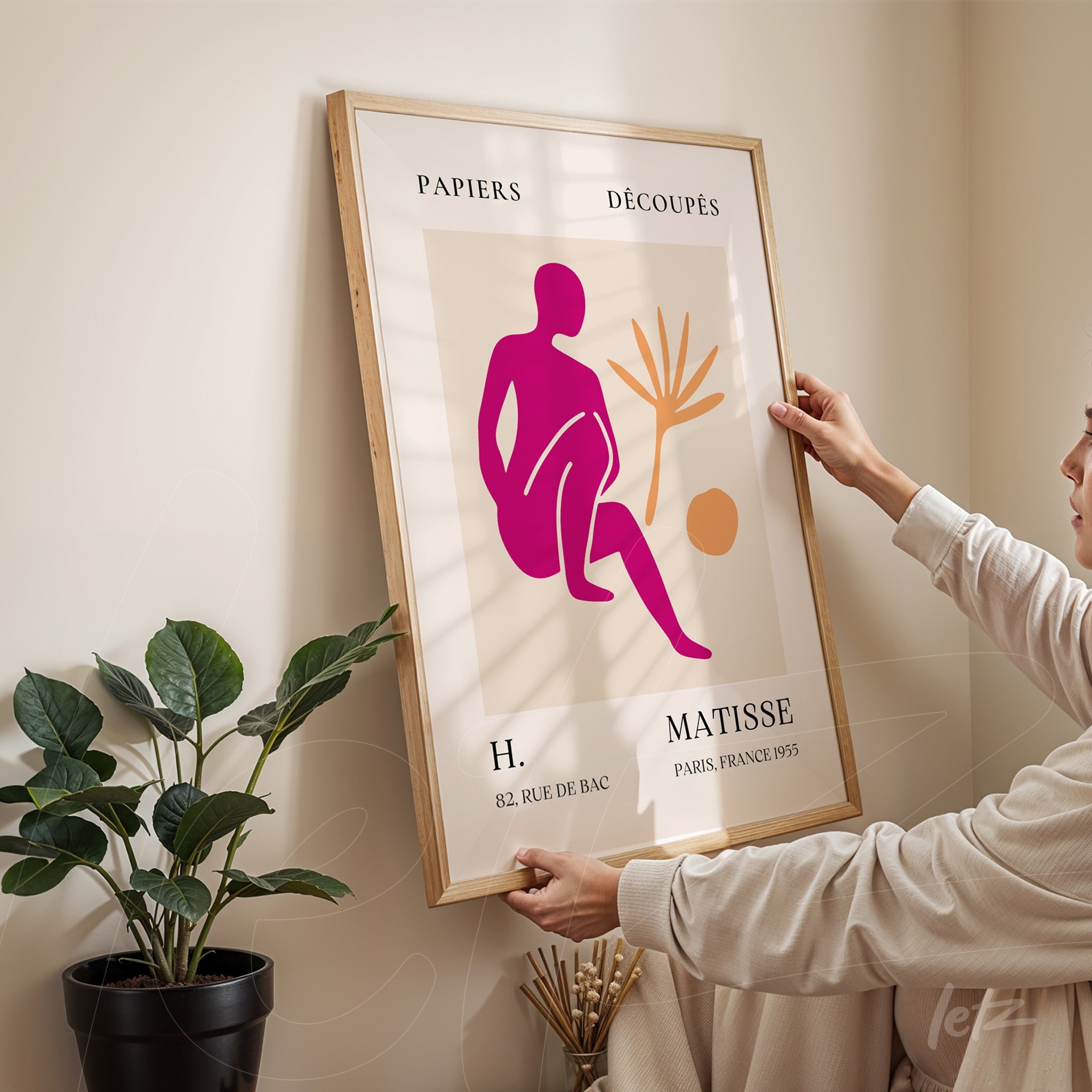 framed decorative art piece featuring an abstract figure in pink and orange tones, being adjusted by a person in a softly lit indoor setting with a light wood frame