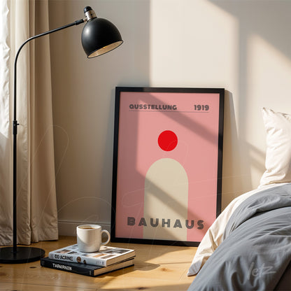 Bauhaus style poster print with simple geometric shapes in a black frame resting on the floor next to a bed and floor lamp