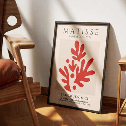 framed modern art print in red tones against a neutral background, leaned beside a wooden chair in a bright room
