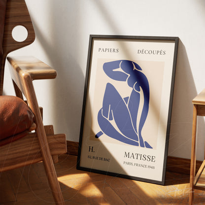 framed artwork in minimalist style featuring a blue female figure, leaning against a light wall next to a modern chair