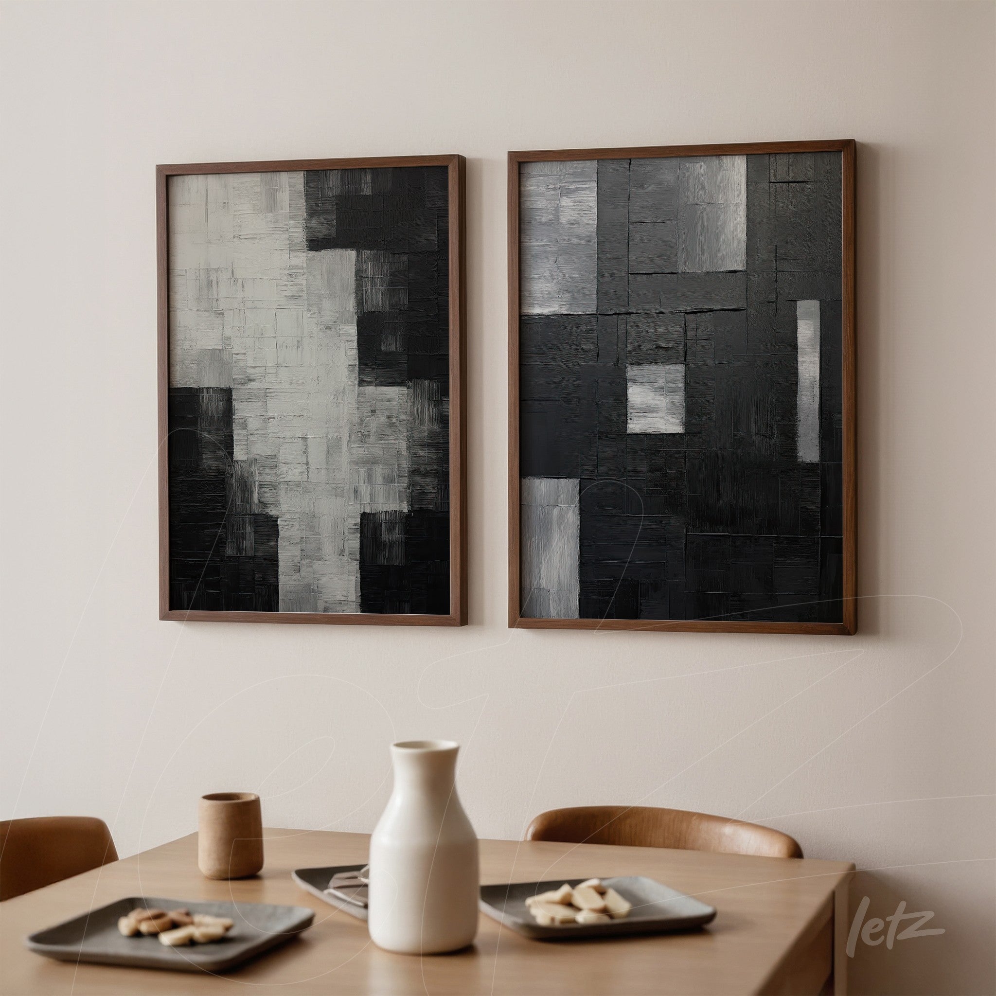 composition featuring two abstract wall art pieces in shades of black and gray, framed in light wood, displayed on a neutral wall above a light wood table