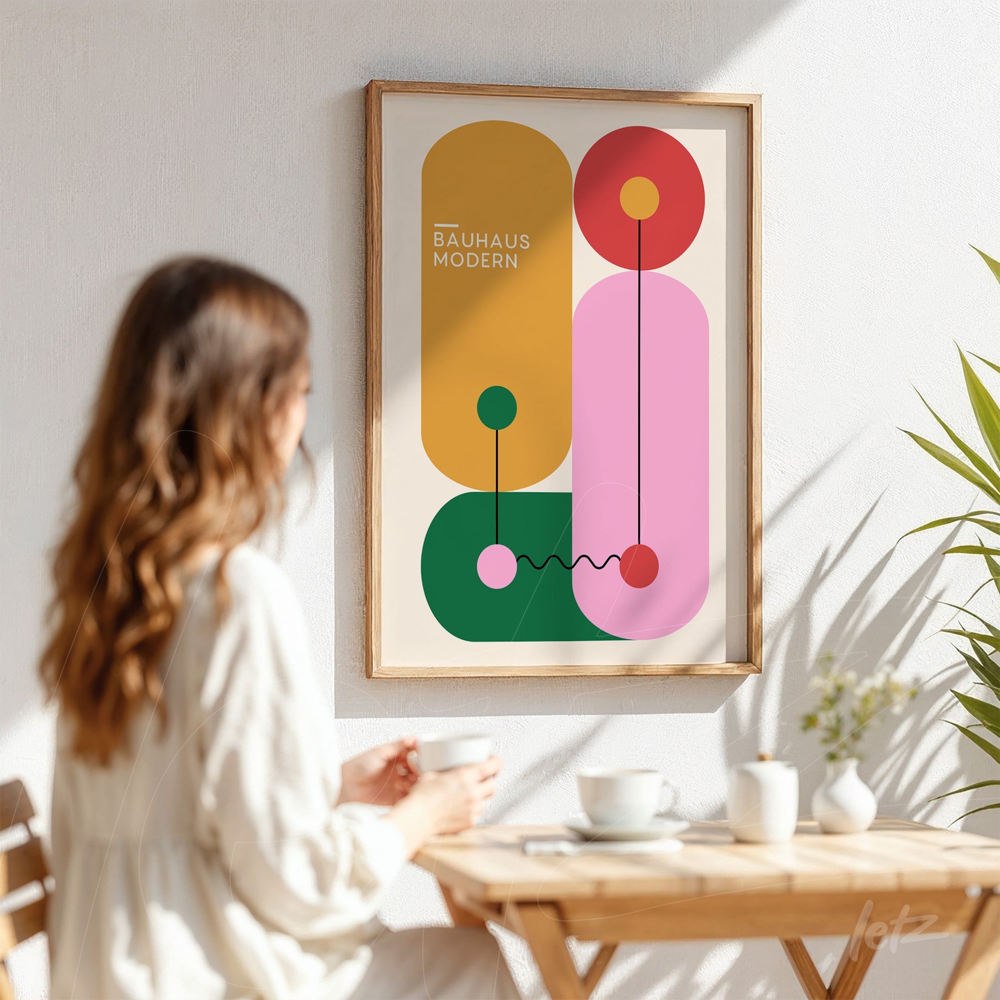 poster print featuring colorful geometric bauhaus-style design in a light wood frame, displayed on a white wall next to a table with cups