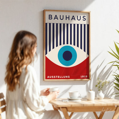 poster print featuring Bauhaus style art with a central blue eye, vertical stripes and light wood frame displayed on a white wall next to a seated woman