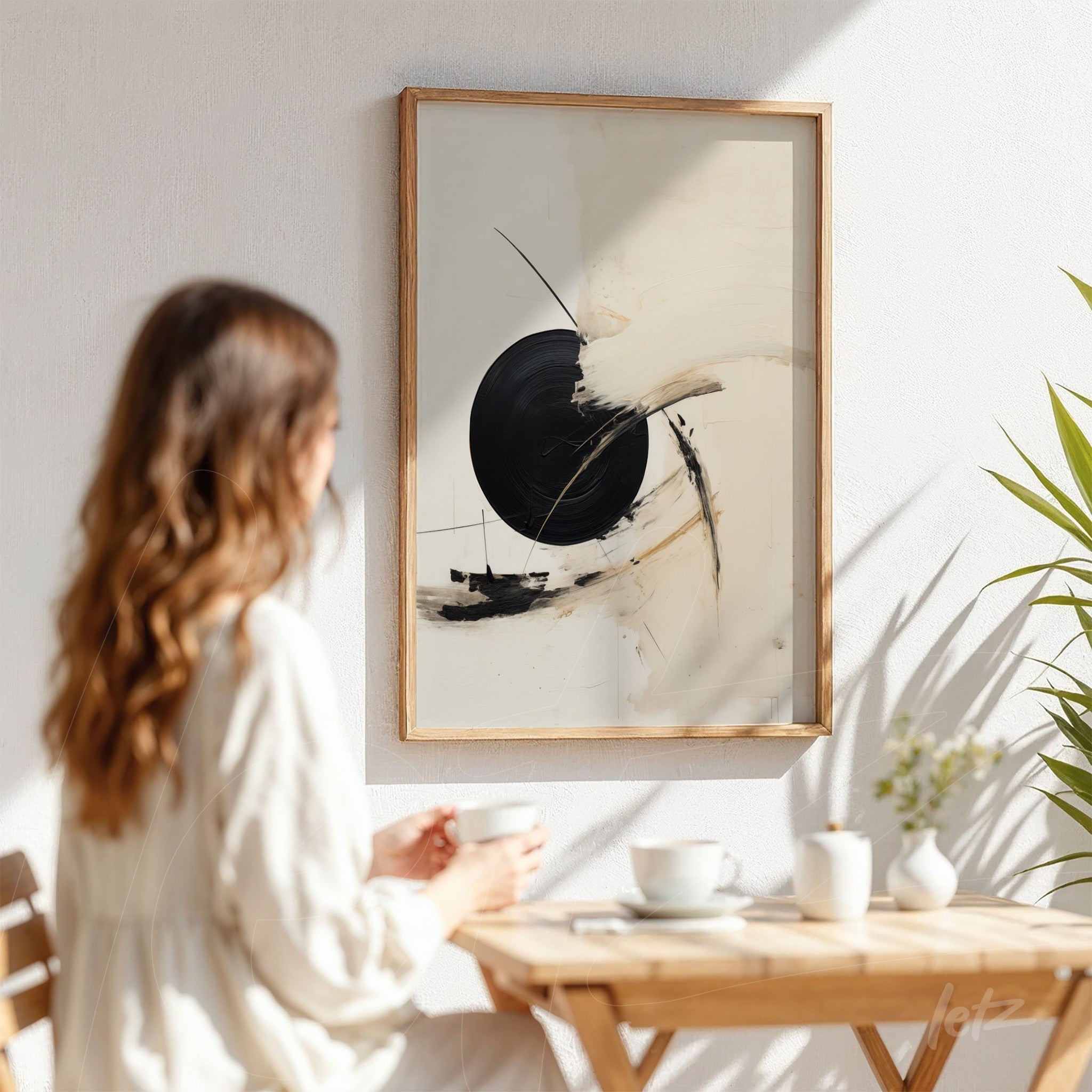 abstract artwork featuring a black and white composition in a light wood frame displayed on a white wall with a woman sitting at a wooden table