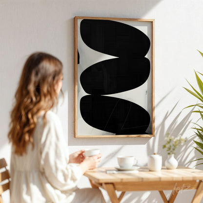 abstract wall art featuring black shapes on an ivory background, framed in light wood, displayed on a light wall next to a green plant