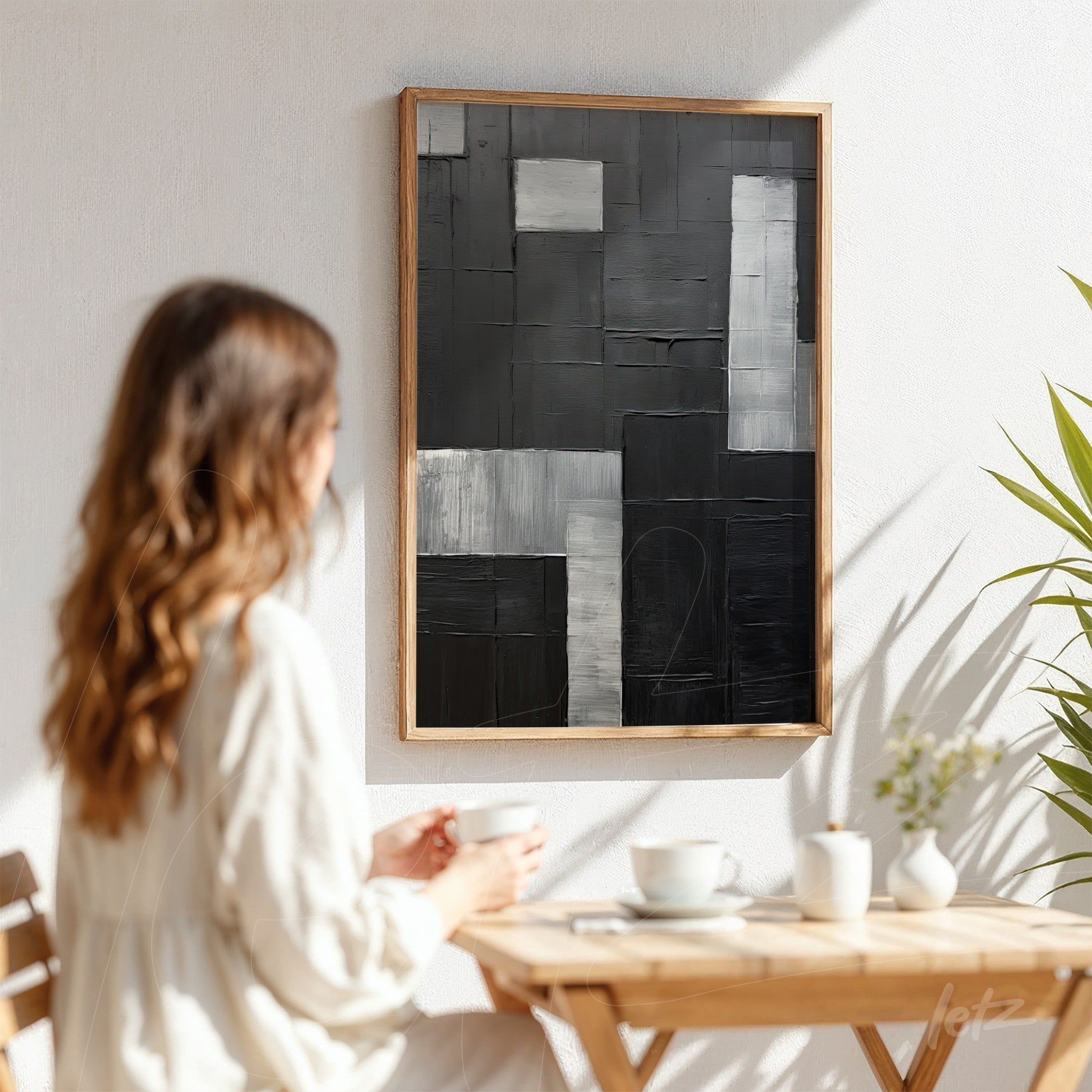 abstract artwork in shades of black and gray with light wood frame displayed on white wall with a woman sitting and admiring the art