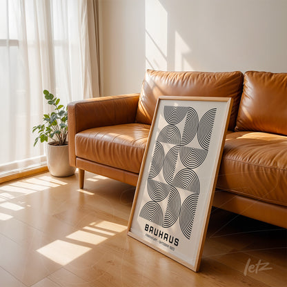 poster art with geometric Bauhaus-inspired design leaning against a brown sofa in a minimalist room with light wood flooring