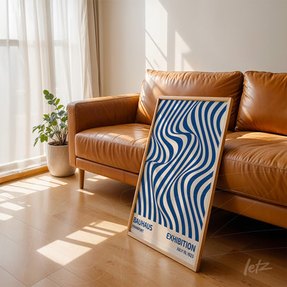 poster featuring wavy blue abstract art framed in light wood, leaning against a brown leather sofa in a sunlit room