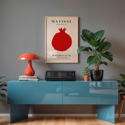 framed artwork by Matisse featuring a red pomegranate on light background displayed above a glossy blue table with an orange lamp and a potted plant