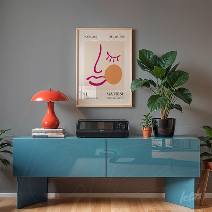 framed modern art print featuring a stylized face in gold frame above a blue table, accompanied by a red lamp and plant