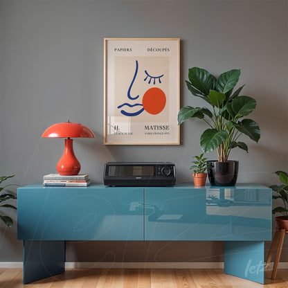 framed wall art in gold frame featuring a minimalist illustration of a face with red accents displayed on a blue table with a potted plant beside it