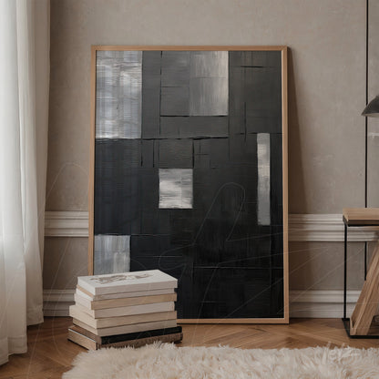 canvas artwork featuring abstract design in shades of black and gray, framed in light wood, displayed on a table beside a stack of books
