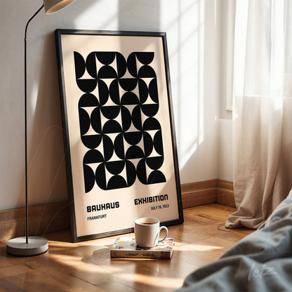 poster featuring black geometric design on beige background in black frame leaning against a white bedroom wall with lamp and cup on the floor