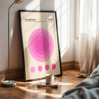 poster print with concentric pink circles on beige background, propped on wooden floor with black frame, natural light on the left
