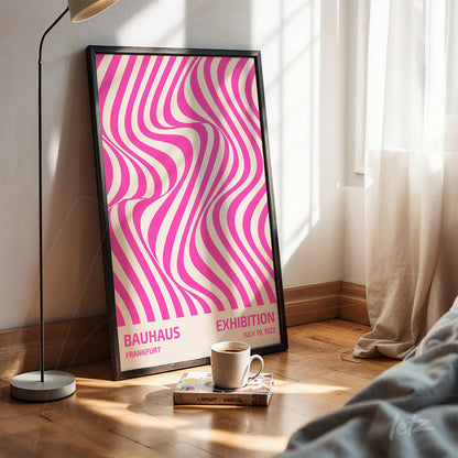 poster with abstract pink and white wave design in black frame, resting on wooden floor next to a cup and book