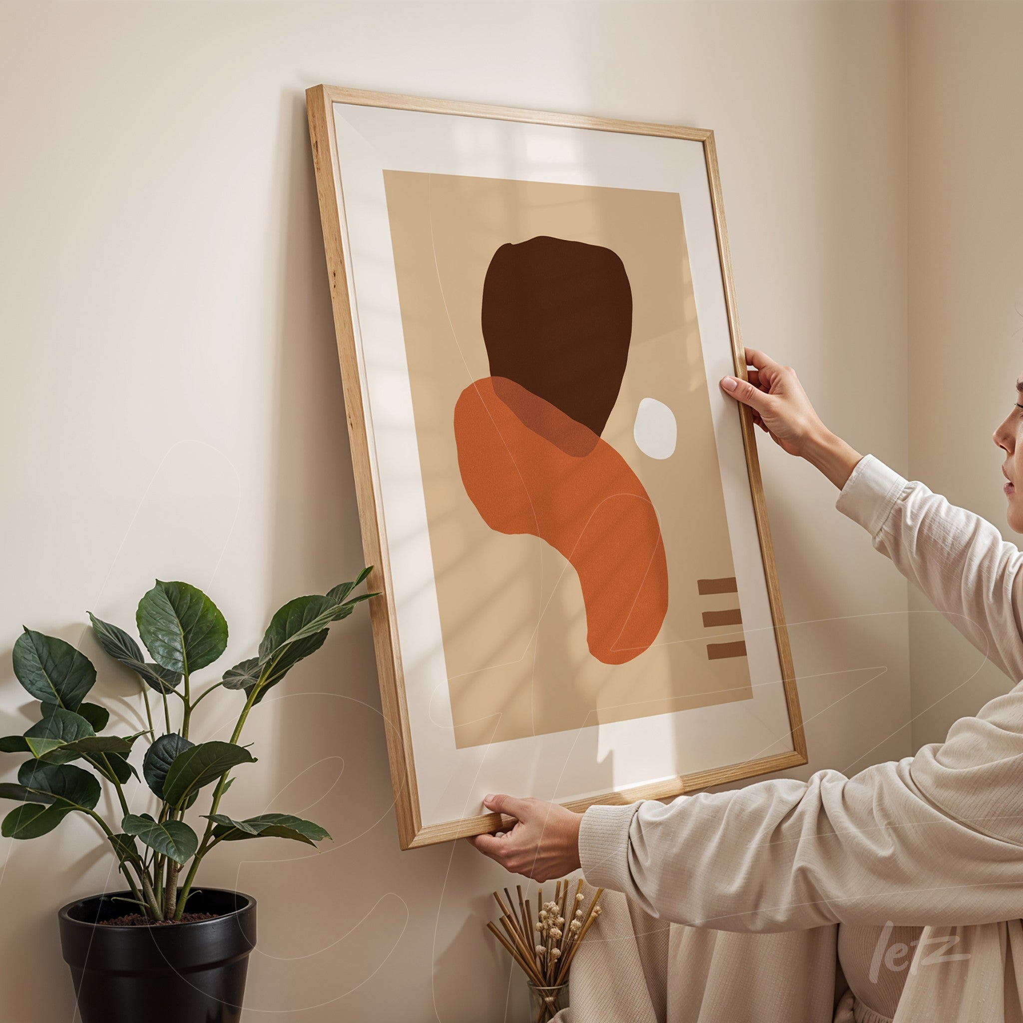 abstract artwork in earthy tones with organic shapes in a light wood frame, held by a person against a cream wall next to a green plant in a black pot