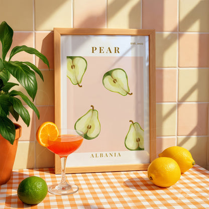 framed wall art featuring stylized green pears on a light wooden frame, placed on an orange and white checkered tablecloth alongside a cocktail glass and fruits