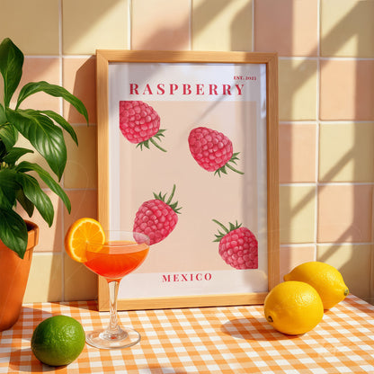framed artwork featuring raspberry illustrations on a light background, with a light wood frame, accompanied by a cocktail glass and citrus fruits on a checkered table