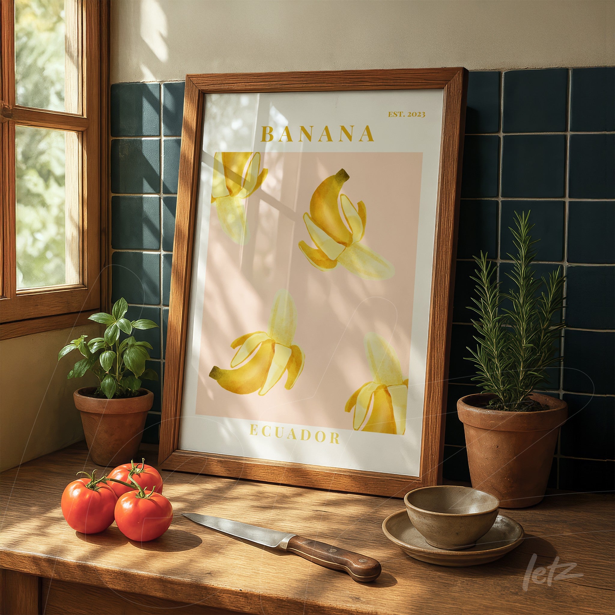 framed artwork featuring banana illustration on pink background displayed on wooden table next to potted plants and tomatoes