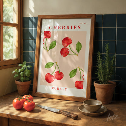 framed artwork of cherries on pink background with light wood frame displayed on a wooden table alongside red tomatoes and a knife