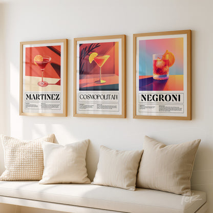 composition of three framed artworks in gold frames featuring colorful cocktail illustrations of martinez, cosmopolitan, and negroni on a light wall