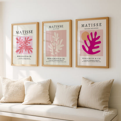 composition of three framed artworks in beige frames featuring abstract designs in shades of pink and red displayed on a light wall with neutral cushions on a fabric sofa