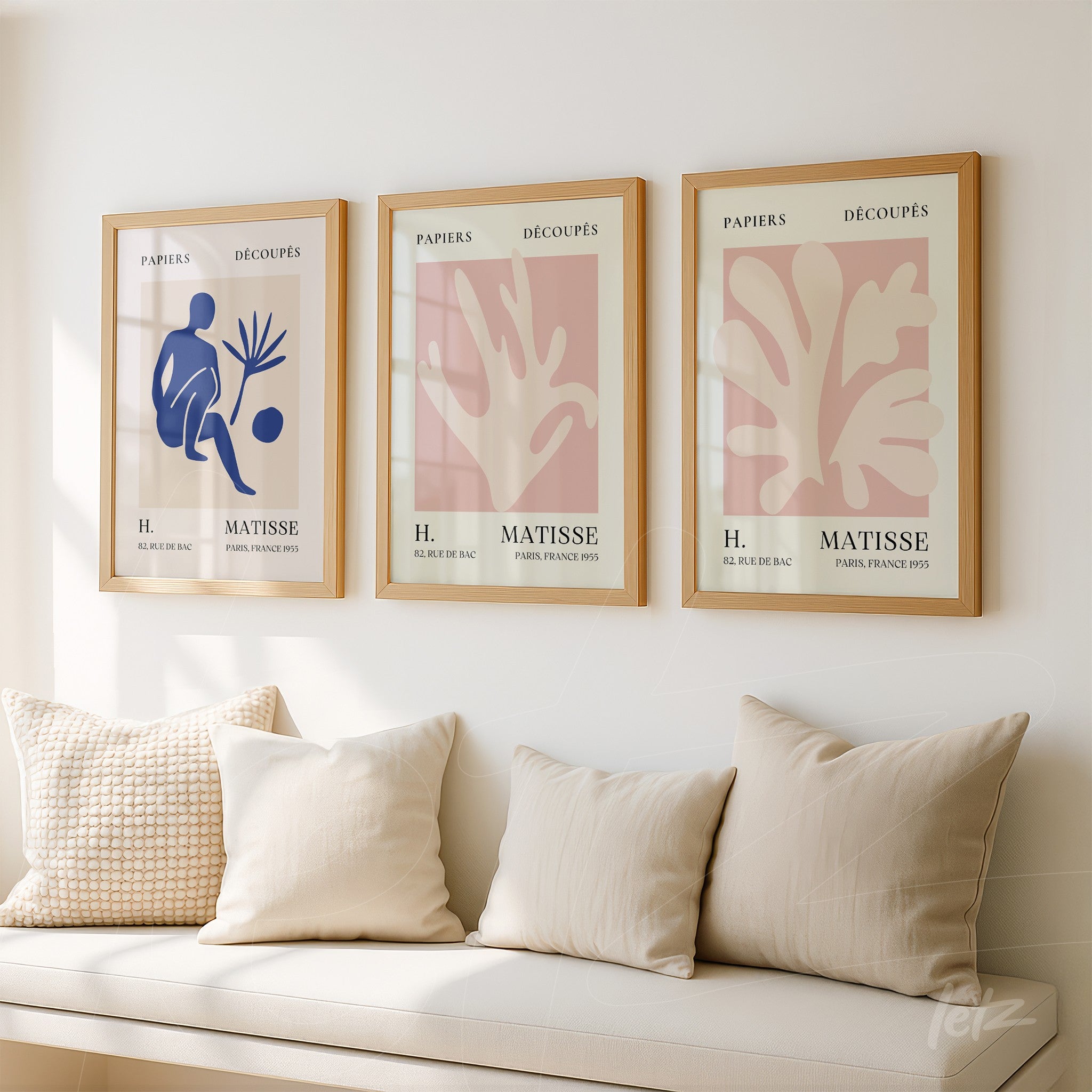 three framed wall art pieces featuring paper cutouts of abstract plant shapes in shades of blue and pink, displayed on a light wall above beige cushions