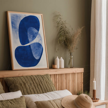 framed abstract art in blue on light background displayed in a room with wooden headboard and natural elements