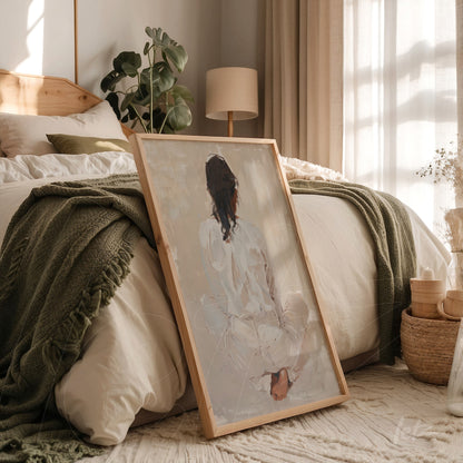 framed artwork of a sitting female figure in a light wooden frame leaning against a bed with neutral bedding in a naturally lit room
