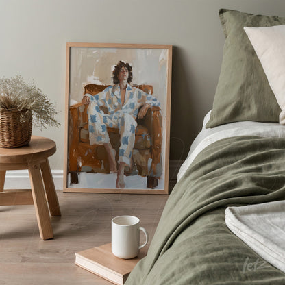 framed artwork in light wood featuring a portrait of a person relaxing on a sofa, dressed in blue pajamas with white patterns next to a bed with neutral-toned bedding