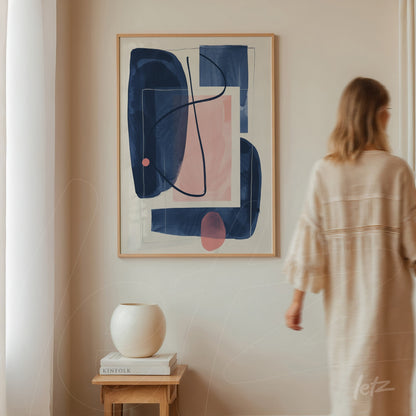 framed abstract artwork featuring geometric shapes in blue and pink displayed on a neutral wall with a woman walking in the background