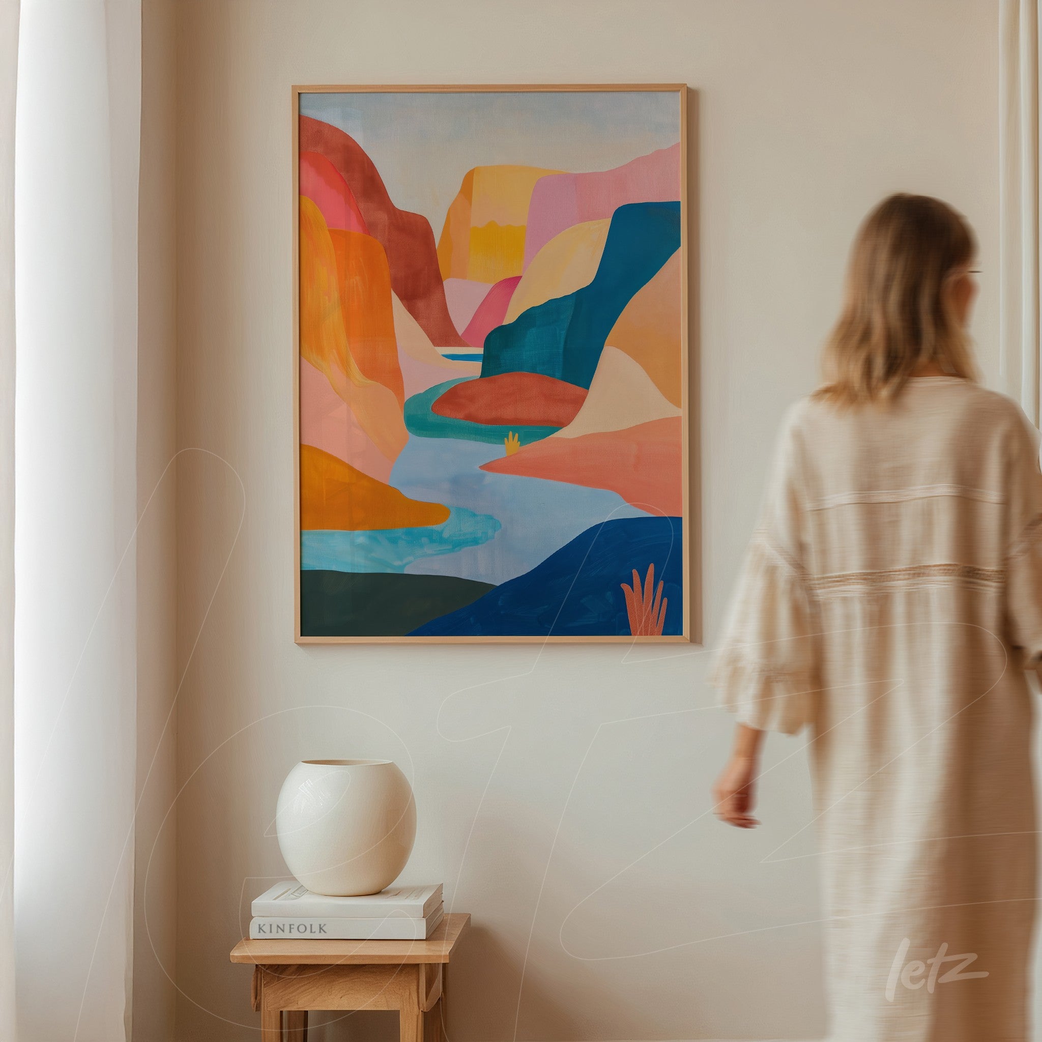 framed abstract art of colorful mountain landscape with light wood frame displayed on a light wall featuring a person walking in the background