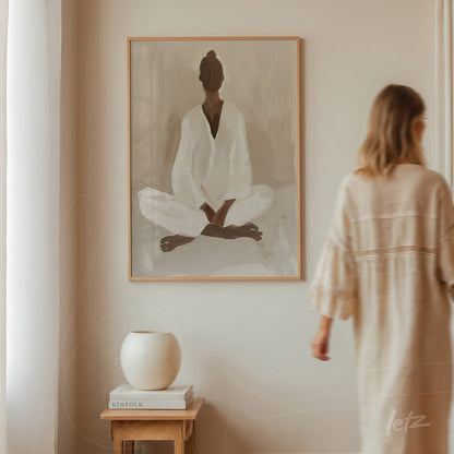 framed abstract art of a seated figure in meditation with a light wood frame displayed on a neutral wall beside a vase and books on a wooden table