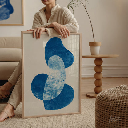framed wall art featuring abstract blue shapes held by a woman in a sweater next to a wooden table